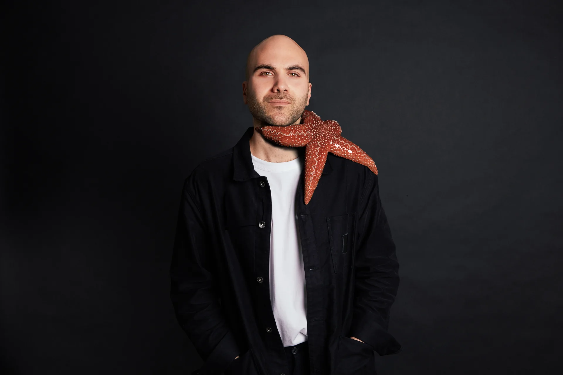 Man with a red starfish on his neck, posing in a black jacket in front of a dark studio background