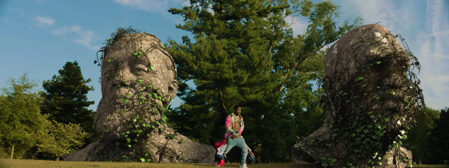 Music video visual featuring stylized 3D stone head covered in vines, with artist performing in a natural setting, part of Marco Mori’s animated music video collection